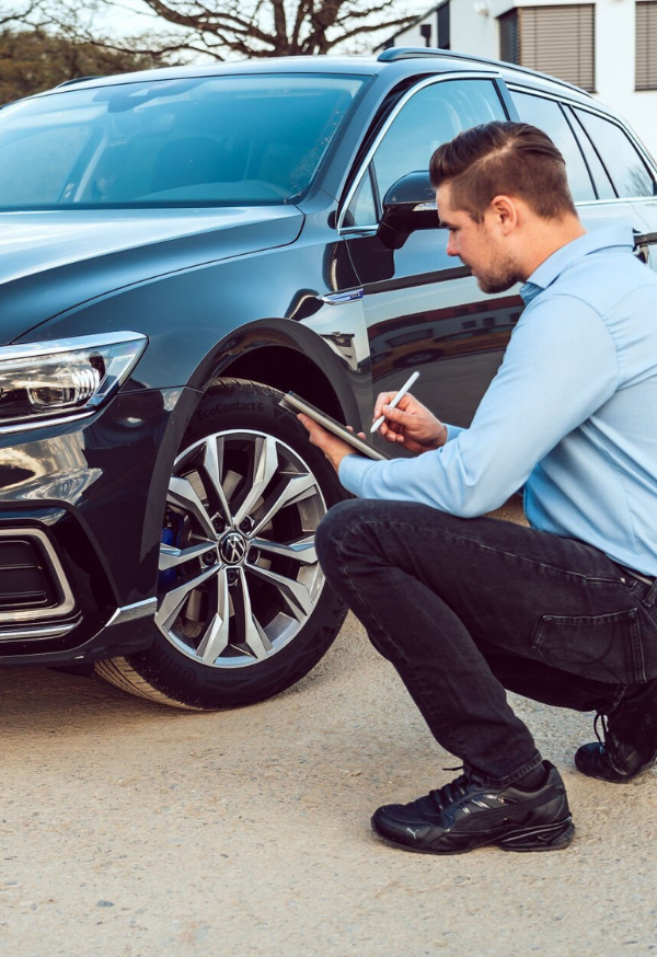 alt="Kfz-Gutachter dokumentiert Fahrzeugschaden vor Ort – Teil der praktischen Ausbildung zum Sachverständigen" Ein Mann überprüft das Rad eines schwarzen Autos und macht Notizen.